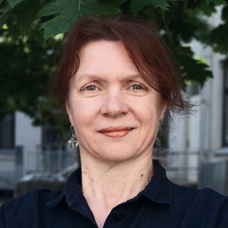 Dr. Berita Simonaitienė pozuoja žalios augmenijos fone, dėvi tamsius drabužius, rausvai rudi plaukai, švelni išraiška.