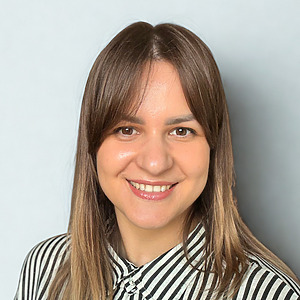 Kristina &Scaron;maižienė &scaron;ypsosi prie&scaron;ais &scaron;viesų foną, rudi plaukai ties pečiais, vilki baltą ir juodą dryžuotą palaidinę.