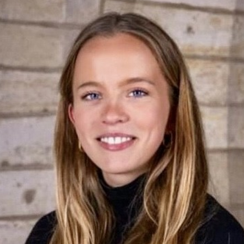 Dr. Simona Sankalaitė šypsosi priešais rusvo akmens sieną, ilgi šviesūs plaukai, vilki juodą megztinį aukštu kaklu.