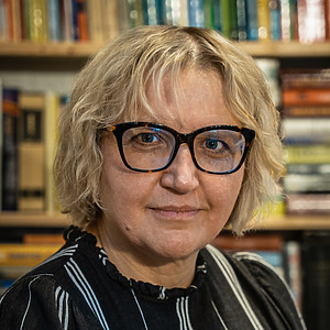 Dr. Vilija Dabri&scaron;ienė fotografuota bibliotekos fone, trumpi &scaron;viesūs plaukai, akiniai su tamsiais rėmeliais, dėvi juodą dryžuotą palaidinę.