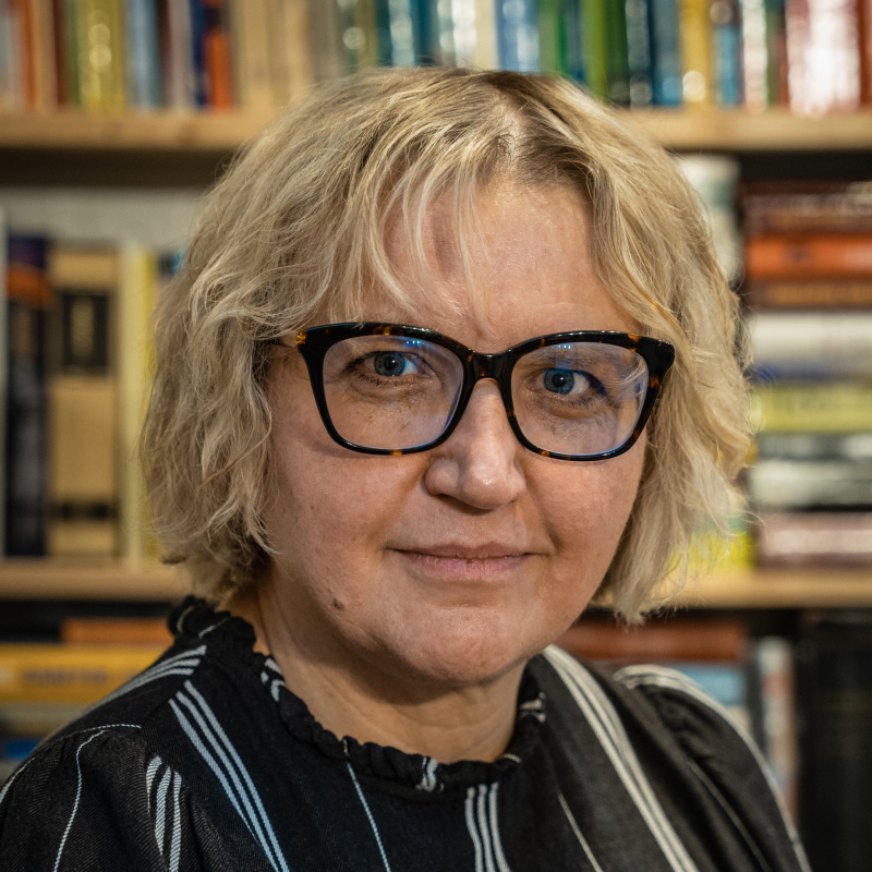 Dr. Vilija Dabrišienė fotografuota bibliotekos fone, trumpi šviesūs plaukai, akiniai su tamsiais rėmeliais, dėvi juodą dryžuotą palaidinę.