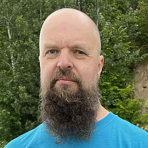 Dr. Giedrius Žvliauskas stovi lauke žalios gamtos fone, trumpai kirptas vir&scaron;ugalvis, ilga ve&scaron;li barzda, vilki mėlynus mar&scaron;kinėlius.